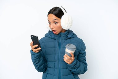 Young hispanic woman wearing a winter earmuffs isolated on white background holding coffee to take away and a mobile