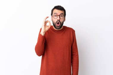 Sakallı, beyaz arka planda beyaz, beyaz, beyaz, yakışıklı bir adam şaşırmış ve iyi bir işaret gösteriyor.