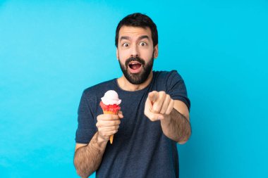 Kornet dondurması ile izole edilmiş mavi arka planı olan genç adam şaşırmış ve ön tarafı işaret ediyor.