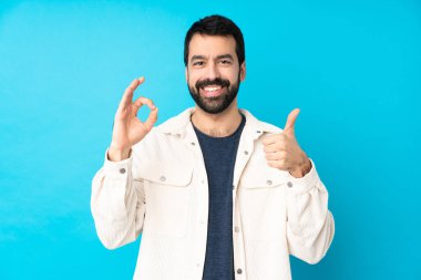 İzole edilmiş mavi arka planda beyaz kadife ceketli genç yakışıklı adam işaret ve baş parmak işareti gösteriyor.