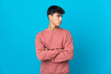 Soyutlanmış mavi arka plandaki genç adam kollarını çapraz tutuyor.