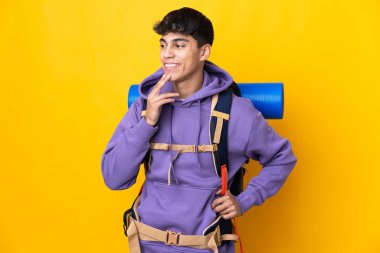 Sırtında büyük bir sırt çantası olan, izole edilmiş sarı arka planı olan genç dağcı gülümserken yukarı bakıyor.