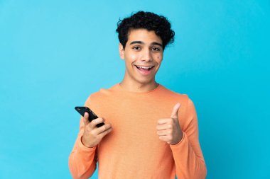 Venezüellalı adam mavi arka planda başparmaklarını kaldırırken cep telefonu kullanıyor.