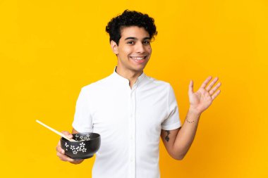 Genç Venezuelalı bir adam sarı arka planda izole edilmiş, elinde çubuklarla elinde makarna tutarken mutlu bir ifadeyle selam veriyor.