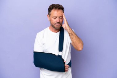 Kırık kollu orta yaşlı beyaz bir adam ve mor arka planda başağrısı olan bir askı takıyor.