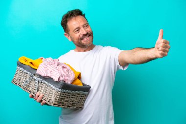 Mavi arka planda izole edilmiş çamaşır sepetini tutan orta yaşlı bir kadın baş parmak işareti yapıyor.