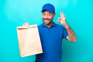 Orta yaşlı bir adam mavi arka planda izole edilmiş bir paket yiyecek alıyor. Parmaklarıyla tamam işareti gösteriyor.