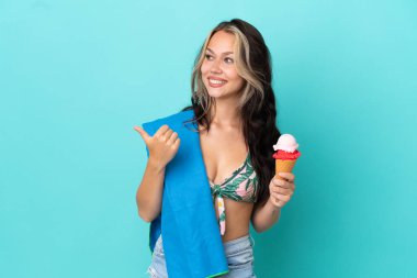 Elinde dondurma ve havluyla mavi arka planda izole edilmiş beyaz kız bir ürünü sunmak için yan tarafı işaret ediyor.