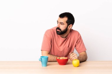Masada kahvaltı yapan beyaz bir adam yan tarafa bakıyor..