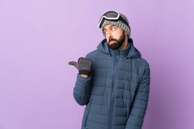 İzole edilmiş mor arka planda snowboard gözlüklü kayakçı adam mutsuz ve bir tarafı işaret ediyor.