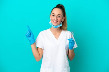 Elinde mavi arka planda izole edilmiş aletler tutan beyaz dişçi kadın en iyinin işareti olarak parmağını kaldırıyor.