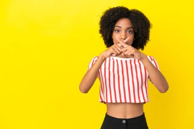 Sarı arka planda izole edilmiş genç Afrikalı Amerikalı kadın sessizlik işareti gösteriyor.