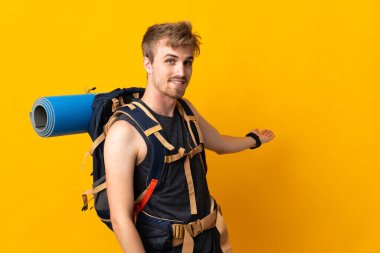 Sarı arka planda izole edilmiş büyük sırt çantalı genç dağcı, davet edildiği için ellerini yana uzatıyor.