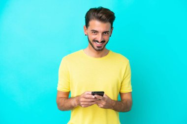 Mavi arka planda izole edilmiş genç beyaz adam cep telefonuyla bir mesaj gönderiyor.