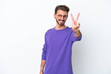 Beyaz arka planda izole edilmiş genç beyaz adam gülümsüyor ve zafer işareti gösteriyor.