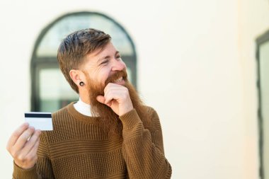 Kızıl saçlı, sakallı bir adam dışarıda kredi kartı tutuyor. Bir fikir üretiyor ve bakıyor.
