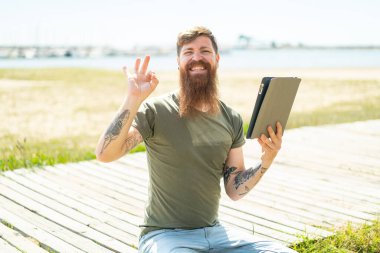 Kızıl saçlı, sakallı bir adam açık havada tablet tutuyor. Parmaklarıyla tamam işareti gösteriyor.