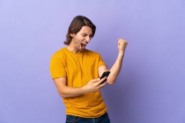 Mor arka planda izole edilmiş, telefon zafer pozisyonunda genç yakışıklı bir adam.