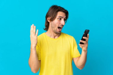 Genç yakışıklı adam mavi arka planda izole edilmiş kameraya bakarken şaşırmış bir ifadeyle cep telefonunu kullanıyor.