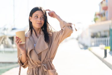 Açık havada kahvesini yudumlayan genç bir kadın kuşkuları ve kafa karıştırıcı bir ifadeyle dışarıda bekliyor.