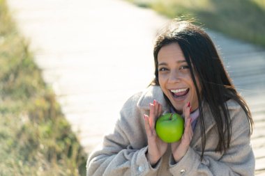 Dışarıda genç bir kadın elinde elmayla şaşkın bir ifadeyle bekliyor.