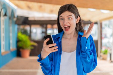 Dışarıda genç ve güzel bir kadın kameraya bakarken şaşırmış bir ifadeyle cep telefonunu kullanıyor.