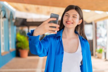 Açık havada genç ve güzel bir kadın cep telefonuyla selfie çekiyor.