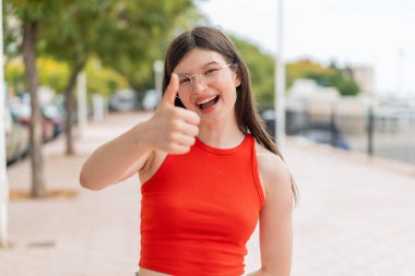 Dışarıda gözlüklü genç bir kadın. Başparmakları havada. Çünkü iyi bir şey oldu.