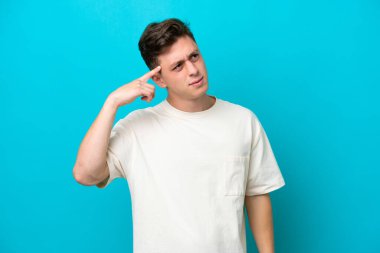 Genç ve yakışıklı Brezilyalı bir adam mavi arka planda izole edilmiş. Delilik jesti yapıyor. Parmağını kafaya koyuyor.
