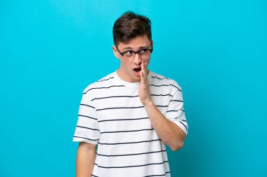 Genç, yakışıklı Brezilyalı bir adam mavi arka planda izole edilmiş. Yan tarafa bakarken sürpriz bir jest yaparak bir şeyler fısıldıyor.