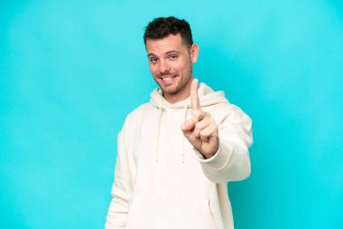 Genç, beyaz, yakışıklı bir adam mavi arka planda izole edilmiş ve parmağını kaldırıyor.