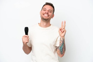 Beyaz arka planda izole edilmiş bir mikrofonu kaldıran genç beyaz şarkıcı gülümsüyor ve zafer işareti gösteriyor.