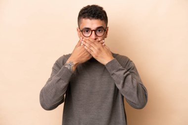 Bej arka planda izole edilmiş genç Brezilyalı adam elleriyle ağzını kapatıyor.