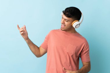 Genç Kolombiyalı adam mavi arka planda izole edilmiş müzik dinliyor ve gitar hareketi yapıyor.