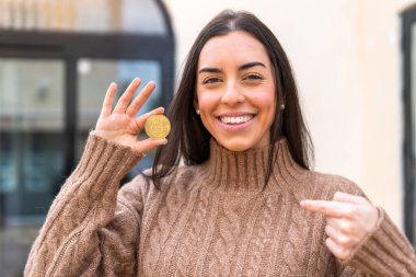 Açık havada elinde Bitcoin tutan genç bir kadın sürpriz bir yüz ifadesiyle.
