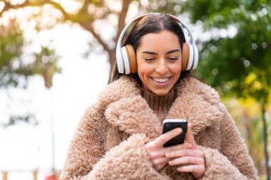 Genç esmer kadın açık havada müzik dinliyor ve cep telefonu arıyor.