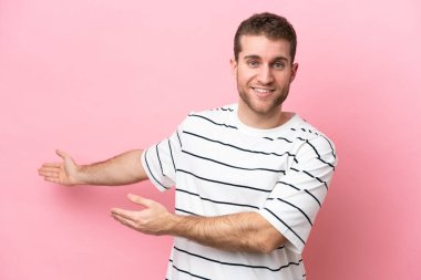 Pembe arka planda izole edilmiş genç beyaz adam, davet edilmek için ellerini yana uzatıyor.