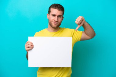 Kafkasyalı genç adam mavi arka planda izole edilmiş. Elinde boş bir levha var ve kötü sinyal veriyor.