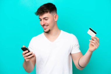 Kafkasyalı yakışıklı genç adam mavi arka planda izole edilmiş ve kredi kartıyla cep telefonuyla alışveriş yapıyor.