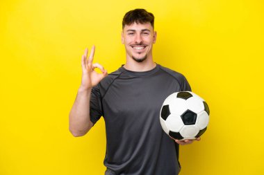 Sarı arka planda izole edilmiş genç futbolcu parmaklarıyla tamam işareti gösteriyor