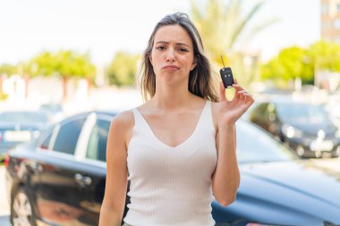 Genç ve güzel bir kadın dışarıda arabanın anahtarlarını tutuyor, üzgün bir ifadeyle.