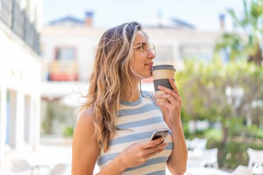 Dışarıda cep telefonu kullanan ve elinde kahve tutan genç ve güzel bir kadın.