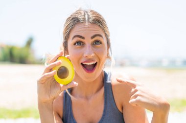 Avokado tutan genç sarışın kadın sürpriz bir yüz ifadesiyle dışarıda.