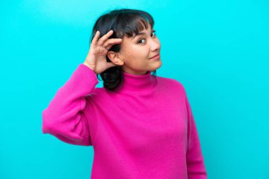 Arjantinli genç bir kadın mavi arka planda izole edilmiş bir şekilde elini kulağına koyarak bir şeyler dinliyor.