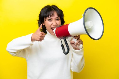 Arjantinli genç bir kadın sarı arka planda bir şey duyurmak için megafonla bağırıyor ve başparmağını kaldırıyor.