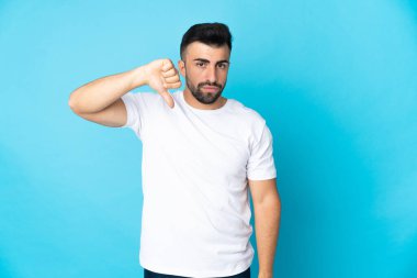 İzole edilmiş mavi arkaplan üzerinde beyaz bir adam negatif ifadeyle baş parmağını aşağı gösteriyor.