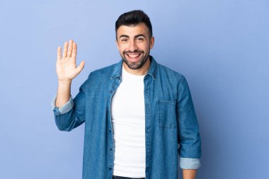 İzole edilmiş mavi arka planda beyaz bir adam mutlu bir ifadeyle selamlıyor.