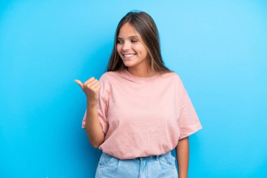 Mavi arka planda izole edilmiş genç beyaz kadın bir ürünü sunmak için kenarı işaret ediyor.