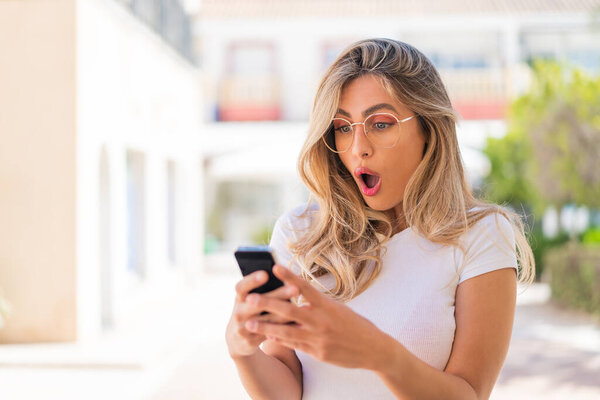 Красивая блондинка Уругвая смотрит в камеру, используя мобильный телефон с удивленным выражением лица