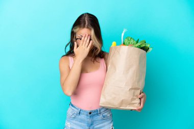Elinde alışveriş torbası tutan genç Rus kadın mavi arka planda izole edilmiş, yorgun ve hasta bir ifadeyle.